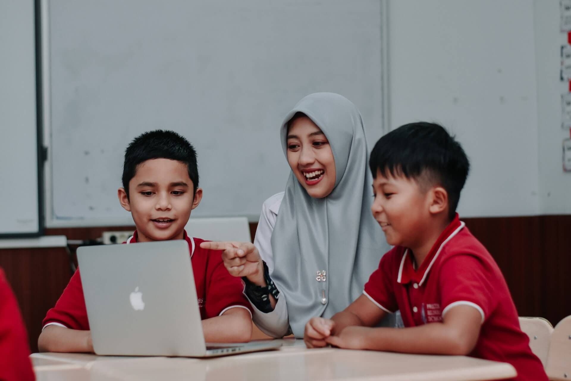 Guru mendampingi dua siswa yang belajar dengan laptop